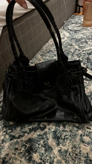 Svart handväska från Paris Hilton - Cool svart handväska från Paris Hilton i skinn med vita kontrastsömmar och detaljer i guld. Väskan har dubbla handtag, dekorativa spännen och dragkedja på insidan. Perfekt storlek för allt du behöver och snygg att bära över axeln.