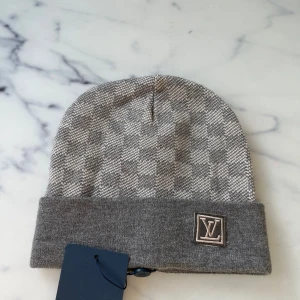  grå mössa - Snygg mössa i grått  med klassiskt rutigt mönster. Mössan har en uppvikt kant med logga framtill och är stickad i mjukt material som känns lyxigt. Perfekt för dig som vill ha en stilren accessoar med designer-vibe.