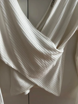 Vit omlottopp med ribbad struktur - Säljer en snygg vit omlottopp från Nelly Trend med ribbad struktur. Toppen har lång ärm och djup v-ringning som ger en cool och modern look. Perfekt att styla med byxor eller kjol