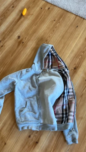 Grå hoodie från Burberry med rutig foder - Säljer en grå hoodie från Burberry i storlek M med klassiskt rutigt foder i huvan och insidan. Tröjan har half zip, huva och en stor magficka. Materialet är mjuk bomull med lite elastan för extra komfort. Perfekt för dig som gillar stilrena och lyxiga detaljer. Dragkedjan krånglar när man ska knäppa den. 