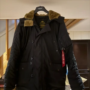 Svart parkajacka Alpha Industries M - Parkajacka från Alpha Industries i svart med fluffig brun fuskpäls vid kragen. Jackan har både dragkedja och knappar framtill, samt flera fickor och en ikonisk röd 'Remove Before Flight'-tag på ärmen. Perfekt för kalla dagar med sin rejäla och snygga look.