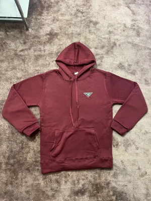 Vinröd hoodie från Prada med huva - Säljer en vinröd hoodie från Prada med klassisk logga på bröstet. Tröjan har huva med snörning, magficka och ribbade muddar vid ärmslut och nederkant. Materialet är mjukt och känns som bomull eller polyester, perfekt för chill dagar.
