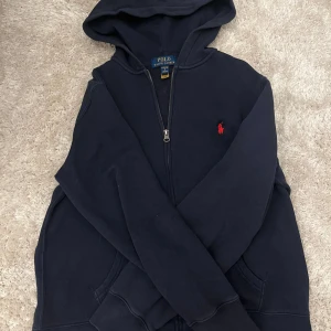 Mörkblå hoodie från Polo Ralph Lauren - Mörkblå hoodie från Polo Ralph Lauren med dragkedja och huva. Klassisk röd logga broderad på bröstet och två fickor framtill. Perfekt för en chill och stilren look. Skön passform och mjukt material.