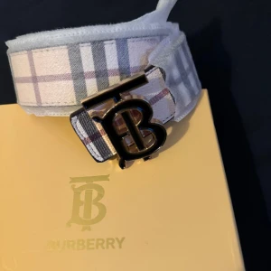 Burberry shoppingväska med klassiskt mönster - Säljer en beige shoppingväska från Burberry med det ikoniska rutig mönstret och en snygg TB-logga i metall. Väskan har rejäla handtag i rep och är tillverkad i slitstark bomull. Perfekt för dig som gillar lyxiga detaljer och klassisk design.