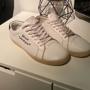 Saint Laurent vita sneakers i skinn - Snygga vita sneakers från Saint Laurent med svart detalj på hälen och handskriven logga på sidan. Skorna har rund tå, platt sula och klassisk snörning. Tillverkade i mjukt skinn med beige yttersula. Perfekta för dig som gillar stilrena och exklusiva sneakers.