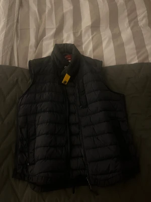 Navy blue dunväst från Parajumpers - Svart quiltad dunväst från Parajumpers med hög krage och gul detalj vid halsen. Västen har dragkedja framtill, sidofickor med dragkedja och en bröstficka. Perfekt för lager-på-lager och har en clean, sportig look. Köpt från Jonhells! Priset går att förhandla