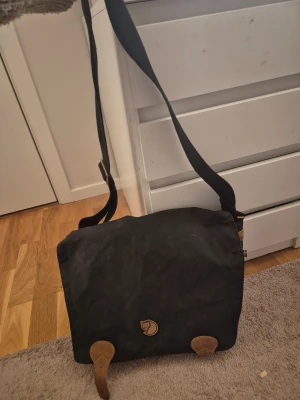 Svart axelväska Fjällräven G-1000 - Svart axelväska från Fjällräven med klassisk G-1000 tyg, bruna skinnremmar och detaljer. Väskan har flera fickor framtill, justerbar axelrem och Fjällrävens logga på locket. Perfekt för dig som vill ha en stilren och praktisk väska med cool outdoor-vibe.