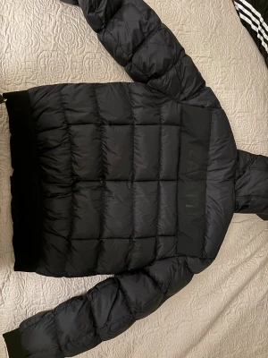 Svart pufferjacka från Zavetti Canada - Svart pufferjacka från Zavetti Canada med huva och dragkedja framtill. Jackan har quiltad design, ribbade muddar och en snygg lönnlövpatch på ärmen. Insidan har tryckt logga och mjukt foder. Perfekt för kalla dagar och riktigt street.