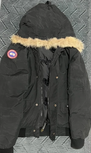 Svart dunjacka från Canada Goose - Säljer en svart dunjacka från Canada Goose med fluffig päls runt huvan och klassisk patch på ärmen. Jackan har ribbade muddar, flera fickor och kraftig dragkedja med logotyp. Perfekt för kalla vinterdagar och riktigt snygg streetstil.
