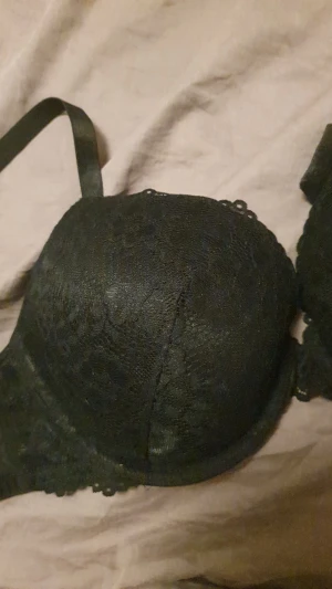 Svart BH I Spets - Säljer en svart bh med vadderade kupor och justerbara axelband. Bhn har ett snyggt spetsmönster över hela ytan och en feminin kant upptill. En liten kudde i upptäckte jag när jag kom hem därav säljer jag.