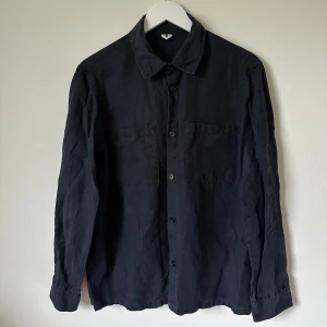 Mörkblå skjortjacka i linne och lyocell - Mörkblå överskjorta med klassisk krage och två stora bröstfickor. Tillverkad i en skön mix av 55% lyocell och 45% linne som ger en lätt och luftig känsla. Långärmad modell med knappar framtill, perfekt för en chill och stilren look. 4Ö18