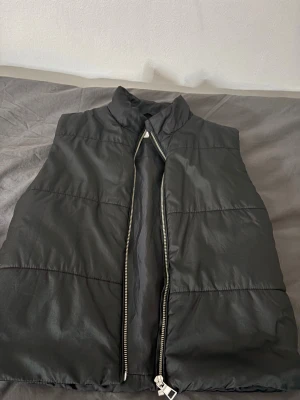 Svart dunväst från KappAhl - Svart dunväst från KappAhl med hög krage och silverfärgad dragkedja framtill. Västen har ett stilrent, quiltat utseende och är tillverkad i syntetmaterial. Perfekt för lager-på-lager och enkel att matcha med din garderob.