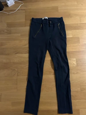 Mörkblå skinny byxor från Hampton Republic - Snygga mörkblå byxor från Hampton Republic med skinny passform. Byxorna har två framfickor med dragkedja i guld och två bakfickor. Materialet är mjukt och stretchigt, perfekt för en avslappnad stil. Passar dig som gillar smalare ben och stilrena detaljer.