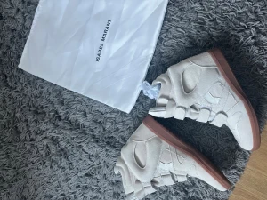 Isabel Marant vita höga sneakers - Säljer ett par snygga höga sneakers från Isabel Marant i ljusbeige mocka med bruna gummisulor. Skorna har rund tå, platt sula och flera kardborreband över foten för en cool look. Perfekt för dig som gillar statement-sneakers med unik design.