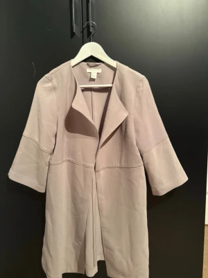 Beige kappa med trekvartsärm - Snygg beige kappa från Wera med öppen front och trekvartsärmar. Jackan har en stilren design med mjukt tyg och slitsar i sidorna. Perfekt för dig som gillar minimalistisk och clean look. Passar dig som vill ha en enkel men trendig jacka.