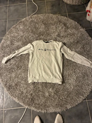 Vit Sail Racing sweatshirt - Säljer en stilren vit sweatshirt från Sail Racing med svart logga tryckt på bröstet. Tröjan har rund halsringning, långa ärmar och ribbade muddar vid ärmslut och nederkant. Perfekt till jeans eller shorts för en clean look. Storlek S ska nog också fungera. Om man har frågor angående tröjan så är det bara att skicka.