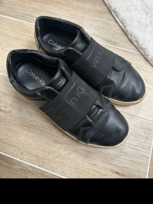 Svarta sneakers från Calvin Klein 38 - Svarta sneakers från Calvin Klein med bred elastisk rem över foten där loggan är tryckt. Skorna har rund tå och platt sula med CK-mönster undertill. Tillverkade i skinn med detaljer i textil. Snygg och stilren design som passar till många olika outfits.