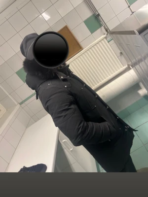 Svart parkajacka från Moose Knuckles - Säljer en svart parkajacka från Moose Knuckles med päls på huvan och snygga metalldetaljer på ärmen. Jackan har flera fickor med knappar och dragkedja, samt en längre passform som håller dig varm under kalla dagar. Perfekt för dig som vill ha en stilren och funktionell vinterjacka. Äkta!