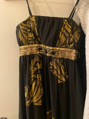 Svart långklänning med guldmönster - Svart långklänning med tunna axelband och detaljerat guldmönster. Midjan pryds av ett brett band med paljetter och svarta stenar som ger extra bling. Tyget är lätt och faller snyggt, perfekt för att sticka ut med en lyxig vibe. Mönstret är inspirerat av barockstil och klänningen har en elegant siluett.