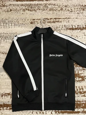 Svart track jacket från Palm Angels - Säljer en svart track jacket från Palm Angels med vita ränder längs ärmarna och vit dragkedja. Jackan har hög krage, två fickor med dragkedja och logga på bröstet. Materialet är slätt och känns sportigt, perfekt för en streetwear-look. Kommer med prislapp använd bara 2 ggr. Jackan är i gott skick men har en liten tråddragking i tyget.