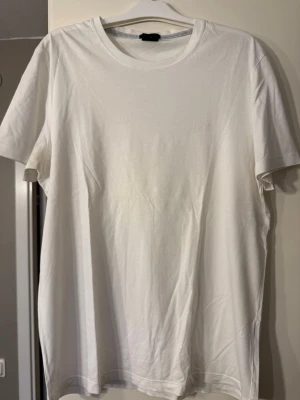 Vit slim fit t-shirt från Hugo Boss - Vit t-shirt från Hugo Boss med diskret broderad logga på bröstet. Modellen är slim fit med rund hals och korta ärmar. Tillverkad i mjuk bomull som känns skön mot huden. Perfekt för dig som gillar stilrena och enkla plagg med premiumkänsla.