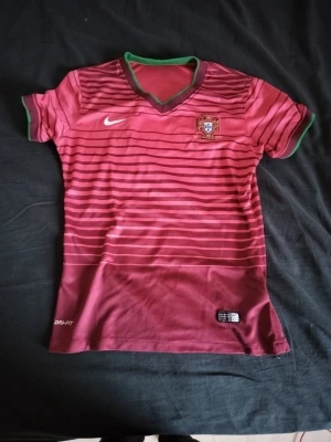 Portugal Nike fotbollströja röd - Säljer en officiell Portugal fotbollströja från Nike med Dri-Fit material. Tröjan är röd med mörkröda horisontella ränder, gröna detaljer vid krage och ärmslut samt broderat Portugal-emblem på bröstet. Perfekt för dig som älskar fotboll och vill ha en snygg landslagströja.