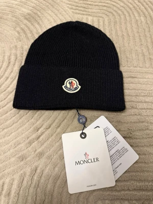 Svart Moncler mössa i ull - Svart ribbstickad mössa från Moncler med klassisk logotyp framtill. Mössan är tillverkad i mjuk ull och har en bred uppvikt kant. Perfekt för kalla dagar och snygg till streetwear. Märkesetikett och QR-kod på insidan.