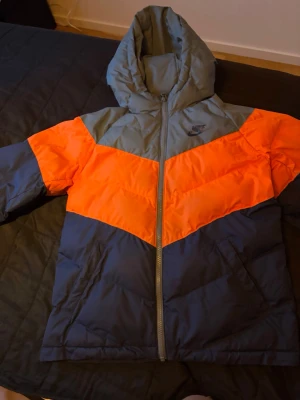 Nike pufferjacka med huva, L - Säljer en snygg pufferjacka från Nike i storlek L 147-158 cm. Jackan har en cool färgblock-design i orange, grått och mörkblått, med huva och dragkedja framtill. Perfekt för kalla dagar och har två sidofickor. Nike-logga på bröstet.