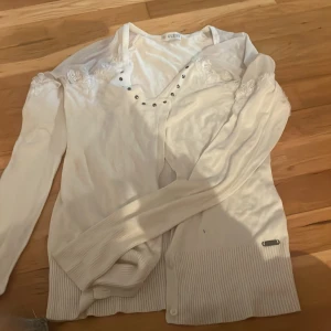 Vit kofta från Guess med spetsdetaljer - Säljer en vit kofta från Guess med snygga spetsbroderier på axlarna och knappar framtill. Koftan har långärmad design och ribbad nederkant. Perfekt för dig som gillar stilrena och feminina detaljer.
