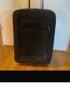 Svart resväska med dragkedja - Stor svart resväska med praktiska dragkedjor och ett rymligt ytterfack. Väskan har en klassisk rektangulär form och är utrustad med hjul och utdragbart handtag för enkel transport. Perfekt för längre resor och packning av mycket kläder och prylar. 55 x 15 x 35 cm 