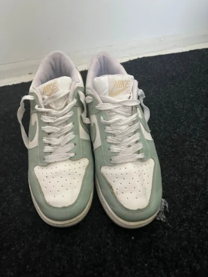 Nike Dunk Low i mintgrön och vit - Nike Dunk Low sneakers i fräsch mintgrön och vit färg med klassisk snörning och rund tå. Ovandelen är i skinn och har perforerade detaljer på tån för extra ventilation. Snyggt broderad Nike-logga på hälen och ikonisk swoosh på sidan.