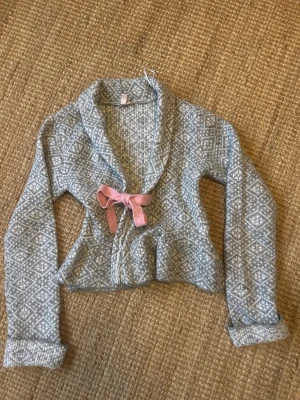 Mönstrad kofta med rosa rosett - Supersöt grå och vit mönstrad kofta med lång ärm och en rosa rosett framtill. Koftan har ett klassiskt stickat mönster och är i mjukt material, perfekt för kyliga dagar. Passar dig som gillar mysiga och unika detaljer.