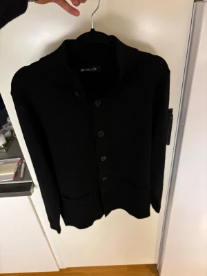 Svart kofta med knappar helt ny! - Svart kofta från Bvaldi med ribbad krage och fem knappar framtill. Koftan har två fickor och är långärmad, perfekt för lager-på-lager. Materialet är stickat och ger en mjuk känsla. Enkel och stilren design som funkar till många outfits. Den är helt sprillans ny aldrig använt bara testat!