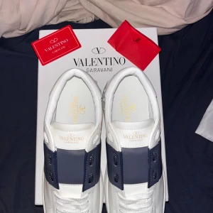 Valentino Garavani Open sneakers vit/blå - Säljer ett par Valentino Garavani Open sneakers i vitt skinn med bred mörkblå rem över snörningen. Snygga detaljer med guldlogga på plösen och innersulan samt mönstrad vit yttersula. Perfekt för dig som gillar lyxiga och stilrena sneakers. (Säljs pga att dom inte passar, köptes på Plick)