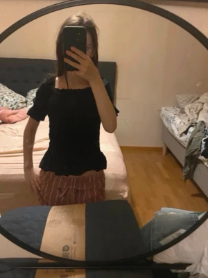 Svart topp med volang - Säljer en svart topp med korta ärmar och volangkant nertill. Toppen har en fyrkantig ringning och sitter tight över kroppen. Perfekt att styla med jeans eller kjol för en trendig look.