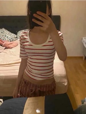 Randig topp - Säljer en croppad, ribbad topp med korta ärmar och rund halsringning. Toppen är vit med röda horisontella ränder och har en figurnära passform. Perfekt för dig som gillar en enkel men snygg stil.