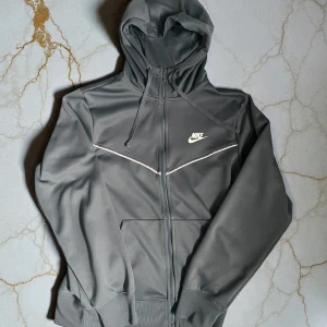 Helt Nike Tech Set - Snygga grå mjukisbyxor från Nike med vit logga på benet och diskreta vita detaljer längs sidorna. Byxorna har elastisk midja och muddar vid bensluten för en skön passform. Perfekta för chill eller träning.