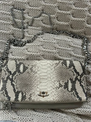 Axelväska i ormskinn från Zadig & Voltaire - Snygg axelväska från Zadig & Voltaire med ormskinnsmönster i beige och grå toner. Väskan har en silverfärgad kedja som axelrem och dragkedja upptill. Märkets logga i metall framtill ger en cool detalj. Perfekt för dig som gillar edgy och trendiga accessoarer.