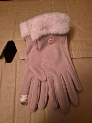 Rosa handskar med mjuk kant upptill - Bilderna visar ett par ljusrosa handskar med mjuk pälsimitation vid mudden och en liten etikett med texten 'Fashion Gloves'. Handskarna har en touchfunktion på ena fingertoppen och är tillverkade i ett tygmaterial.