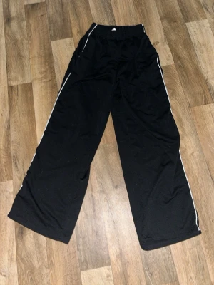 Svarta wide träningsbyxor med vita detaljer - Säljer ett par svarta träningsbyxor med bred passform och vita ränder längs sidorna. Byxorna har elastisk midja och är bekväma. Materialet känns lätt och luftigt, perfekt för dig som gillar loose fit. Från SHEIN och använda ett par gånger