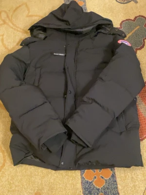 Canada Goose pufferjacka - Svart pufferjacka från Canada Goose med huva och klassisk logga på ärmen. Jackan har quiltad design, långärmade ärmar och dragkedja med knappar framtill. Perfekt för kalla dagar och har en clean, stilren look. Använd ett par gånger fast på ett sparsamt sätt. Skickar inom 24 timmar 💨🚚🙂‍↔️. Vid frågor skriv 💬 