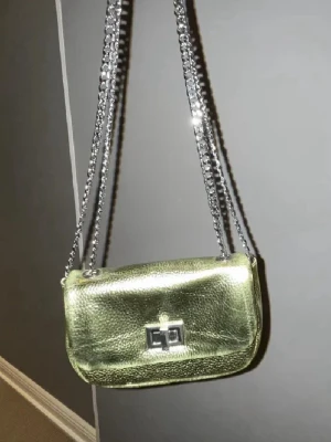 Guldig axelväska med kedja - Säljer en snygg guldig axelväska med glansigt skinnmaterial och silverfärgad kedja som axelrem. Väskan har ett stilrent metallspänne framtill och en kompakt, rektangulär form som gör den enkel att bära med sig. Perfekt för dig som gillar att sticka ut med accessoarer.