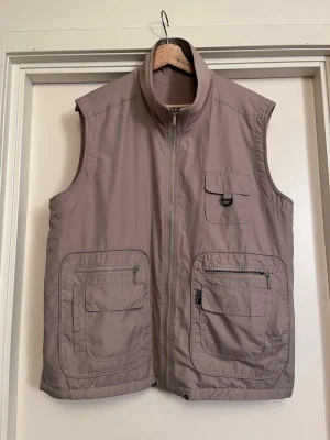 Vändbar utilityväst i fleece  - Vändbar utilityväst i fleece med flera praktiska fickor framtill, både med dragkedja och lock. Västen har hög krage och har fleece invändigt / utvändigt då den är vändbar. Onesize XS-L. Mycket bra skick! Säljs på flera sidor, först till kvarn. 