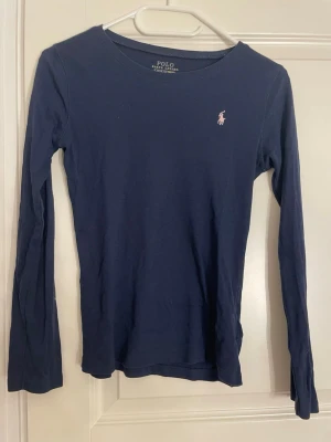 Mörkblå långärmad topp Polo Ralph Lauren - Säljer en mörkblå långärmad topp från Polo Ralph Lauren i mjuk bomull. Toppen har en rund halsringning och en liten rosa broderad logga på bröstet. Perfekt för dig som gillar klassisk och enkel stil med en touch av märke.