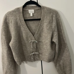 Beige stickad kofta från H&M - Säljer en beige stickad kofta från H&M med v-ringad hals och två snygga knytband framtill. Koftan har långärmade, lite loose ärmar och ribbad struktur som ger en chill vibe. Perfekt att slänga på över en topp när det är lite kyligt ute.