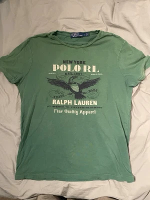 Grön t-shirt från Polo Ralph Lauren - Grön t-shirt från Polo Ralph Lauren med tryck framtill Klassisk rund halsringning och korta ärmar. Stort grafiskt tryck med örn och texten 'New York Polo RL Ralph Lauren  Perfekt för en avslappnad stil.