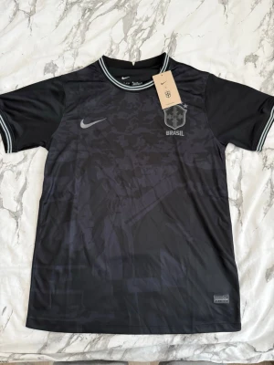 Svart Brasil Nike fotbollströja - Snygg svart Brasil fotbollströja från Nike med diskret kamouflagemönster och silverfärgade detaljer. Tröjan har rund hals med randig kant och Brasil-logga på bröstet. Perfekt för dig som gillar sportig stil och vill visa support för Brasil.