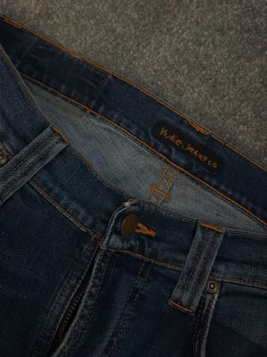 Mörkblå Nudie Jeans Skinny - Säljer ett par mörkblå skinny jeans från Nudie Jeans med klassiska orangea sömmar och snygga broderade bakfickor. Jeansen har fem fickor, dragkedja och knappstängning. Perfekt passform och stilren look för dig som gillar smalare jeans.