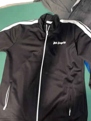 Svart vindjacka från Palm Angels - Palm Angels svart vindjacka med vita detaljer och dragkedja framtill. Jackan har vita ränder längs axlarna och ärmarna, samt logotypen broderad på bröstet. Materialet är polyester och jackan har fickor med dragkedja på sidorna.