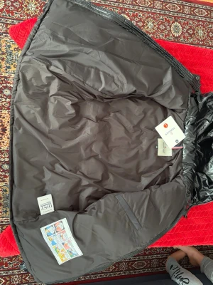 Svart Moncler pufferjacka med huva - Svart pufferjacka från Moncler med glansig finish, huva och dragkedjor på fickorna. Jackan har Moncler-logga på ärmen och quiltad design. Perfekt för kalla dagar och har en riktigt snygg streetstil. Materialet är polyester och dun. Och den går att sckana 